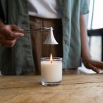 Candle Safety 101: How to Enjoy Your Candles Safely at Home A person snuffs a white candle with a metal tool on a rustic wooden table indoors.