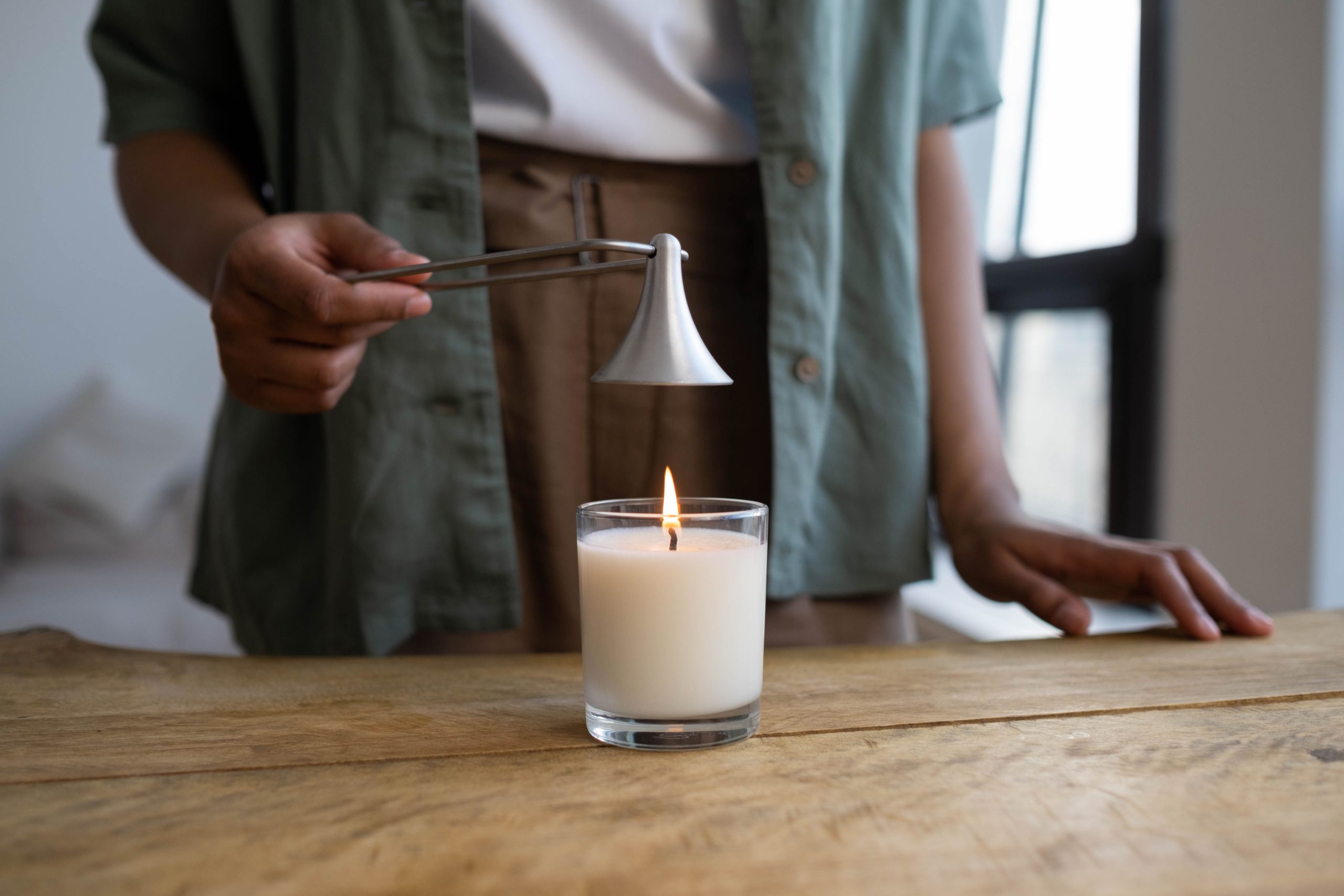 A person snuffs a white candle with a metal tool on a rustic wooden table indoors.